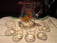 Full set of hand painted glass lemonade pitcher with five matching glasses displayed on a white green-striped cloth. Shows overall set appearance and size.