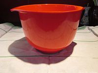 Side profile photo showing the bright orange Rosti mixing bowl with a spout and handle, sitting on a cloth surface.