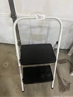 Front view of two-step foldable white metal step stool with black textured steps on concrete floor.
