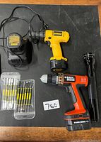 Top view of DeWalt drill, Black & Decker drill, charger, flashlight, and packaged pry tool sets on black surface