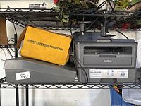 Kodak Carousel Projector in original yellow box placed on top of gray typewriter case, Brother Compact Laser Copier with cables on black metal shelf