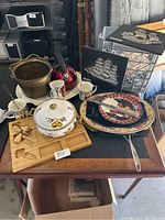 Mixed household items on table including brass bucket, ceramic dinnerware, wooden cutting board, metal decorative plaques, and red kettle.