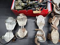 Photo showing four pairs of vintage metal roller skates with leather straps and wheels, along with a box of assorted plastic action figures and a toy vehicle.