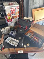 Photo of assorted home items including Black+Decker bag, Mad Chef kit box, battery charger with batteries, small watch, and other miscellaneous items on a table