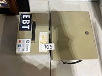 Photograph showing two metal cash lock boxes side by side on a table. The smaller box has various stickers including EBT and Square payment methods on top, black handle, and key lock. The larger box is plain beige with a black handle and key lock.