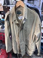 Front view of beige long trench coat hanging on a wooden hanger, showing sleeves, collar, and button details on shoulder straps.