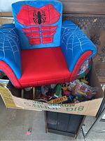 Blue and red Spider-Man themed kids chair with web pattern on armrests, sitting above a box filled with various small toys and Christmas related items.