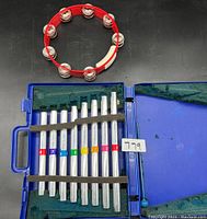 Red tambourine with metal jingles and blue plastic case containing 8 numbered metal chimes inside green foam lining.
