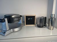 Side view showing Cuisinart waffle maker, Black & Decker food processor, and metal cocktail shaker with Lot 44 sign
