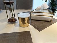 Overview of three items on a white table: tissue holder with brass and glass construction, Eclectic candle in gold container, wood and brass hourglass.