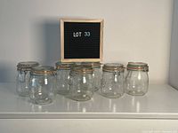 Seven clear glass fermentation jars arranged in two rows with a sign saying LOT 33 behind them. Jars have metal clamp lids and orange rubber seals, knee-high size, round body design.