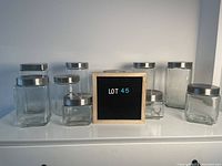 Photo showing assortment of 11 glass jars of round and square shapes with silver metal lids arranged on a white surface.
