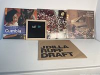 Four international vinyl records displayed, showing cover art and sleeve, including The Rough Guide to Cumbia, crowd photo album, Mondo Grosso Butterfly, and J Dilla Ruff Draft.