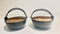 Two black cast iron pots with wooden lids placed side by side on a white surface, showing the tops and handles.