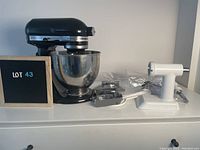 Black KitchenAid Artisan tilt-head stand mixer mounted with stainless steel bowl alongside white plastic pasta press housing and metal pasta roller arm on a white surface.