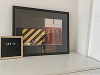 Framed print artwork displayed against a white wall with a lot number sign next to it.