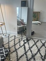 Full-length white mirrored jewelry cabinet standing upright against a wall. Reflects part of the room including a couch and window.