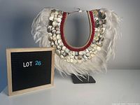 Front view of the neckplate showing feather fringe, shells in layered scallops, and red bead trim, displayed on metal stand.