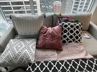 Seven decorative pillows displayed on a beige couch, showing various patterns and colors including white with blue trim, pink, black and white geometric, and multicolor embroidered with pom-poms.