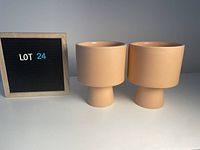 Two blush/adobe colored ceramic planters with cylindrical tops and cone-shaped bases shown next to a sign displaying lot number.