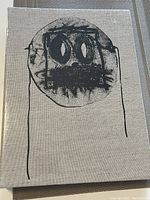 Front cover close-up of the textured beige hardcover book with black abstract face design.