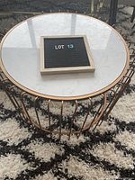 Close-up angled photo showing the white faux marble circular tabletop and rose gold-colored vertical metal rod base on a light patterned rug.