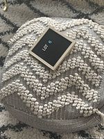 Grey and white soft ottoman with a small sign reading 'LOT 6' on top showing the item's top and part of the surface texture.
