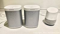 Two white Sonos Play:1 speakers alongside a white Google Home speaker on a table