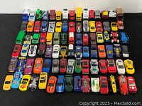 Top-down image of 90 die cast vehicles arranged in rows on a black surface, showing a wide variety of colors and vehicle types including sports cars, trucks, and race cars.