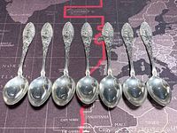 Seven small sterling silver spoons arranged side by side, showing the front and handle detail with embossed floral and leaf design, confirming quantity and condition.