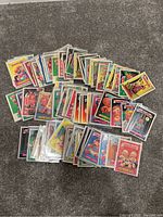 Photo showing approximately 90 Garbage Pail Kids cards fanned out on a carpeted floor. Cards are colorful, feature various characters, and include some sleeved cards.