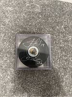 Top view of black hockey puck with Chicago Blackhawks logo and two silver autographs visible, inside clear plastic display case, placed on carpeted floor.