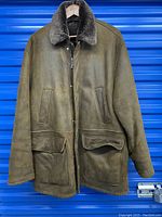 Front view of the brown shearling coat jacket with fur collar and pockets hanging on wooden hanger against blue background.
