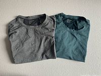 Two folded men's Arc'teryx short sleeve shirts size Large, one grey and one teal, displayed on a light surface.