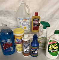 Photo of assorted household cleaning products including ammonia, disinfectant wipes, leather cleaner, multi-surface sprays, and various liquid bottles.