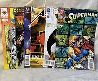Photo showing seven comic books from various publishers, including Valiant, Image, DC Comics, and Marvel. Visible titles include Superman and others with distinct comic book artwork and logos.