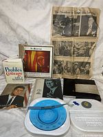 Wide view of the assortment of JFK memorabilia including book, album cover, newspaper page, photo cards, and blue plastic item.