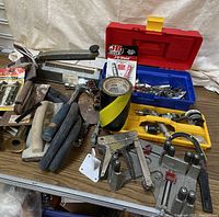 Photo showing assortment of hand tools including stapler guns, clamp, coping saw, sanding block, industrial tape, and containers with hardware and small parts on a table.