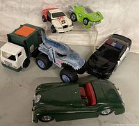 Full lot view showing six toy vehicles on a white cloth background including a garbage truck, monster truck, convertible car, race car, police car, and futuristic car.