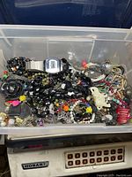 Clear plastic container filled with an assortment of vintage to contemporary broken and unmatched jewelry, beads, and craft findings including chains, a silver digital watch, and various decorative pendants.