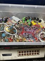 Open plastic bin showing a large mixed collection of jewelry parts including broken necklaces, beads, pendants, and chains in a variety of colors and materials.
