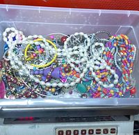 Plastic container filled with assorted broken jewelry pieces including various beads and strands in multiple colors, placed on a digital scale.