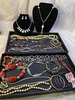 Full overview of assorted vintage, retro, modern and contemporary jewelry pieces displayed on stands and trays. Includes earrings, multiple necklaces, rings, bracelets in various colors and materials.