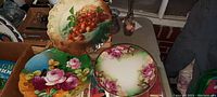 Three antique heavy glass plates displayed on a table, showing colorful floral and fruit designs with scalloped edges.