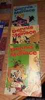 Photo shows three vintage Dennis the Menace comic books stacked vertically on a carpeted floor. Covers show colorful cartoon style artwork with Dennis the Menace character in various scenes. The comic books appear in clean condition with some minor creasing and wear on edges.