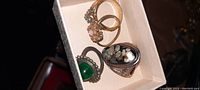 Photo showing four vintage rings inside a white box, including a green cabochon ring, gold-tone rings with clear and opal-like stones, and a silver-tone abalone mosaic ring.