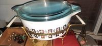 Side view of Pyrex serving dish with clear glass lid on metal stand showing brown leaf pattern and white handles on stand.