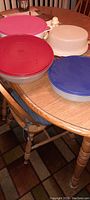Four large round Tupperware containers with different colored lids on a wood table.