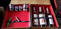 Full inside view of portable bar case containing four empty vintage whiskey bottles, four metal shot cups, a corkscrew, bottle opener, and metal pour spout attachment