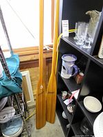 Pair of wooden kayak oars standing upright next to shelving unit with various items.